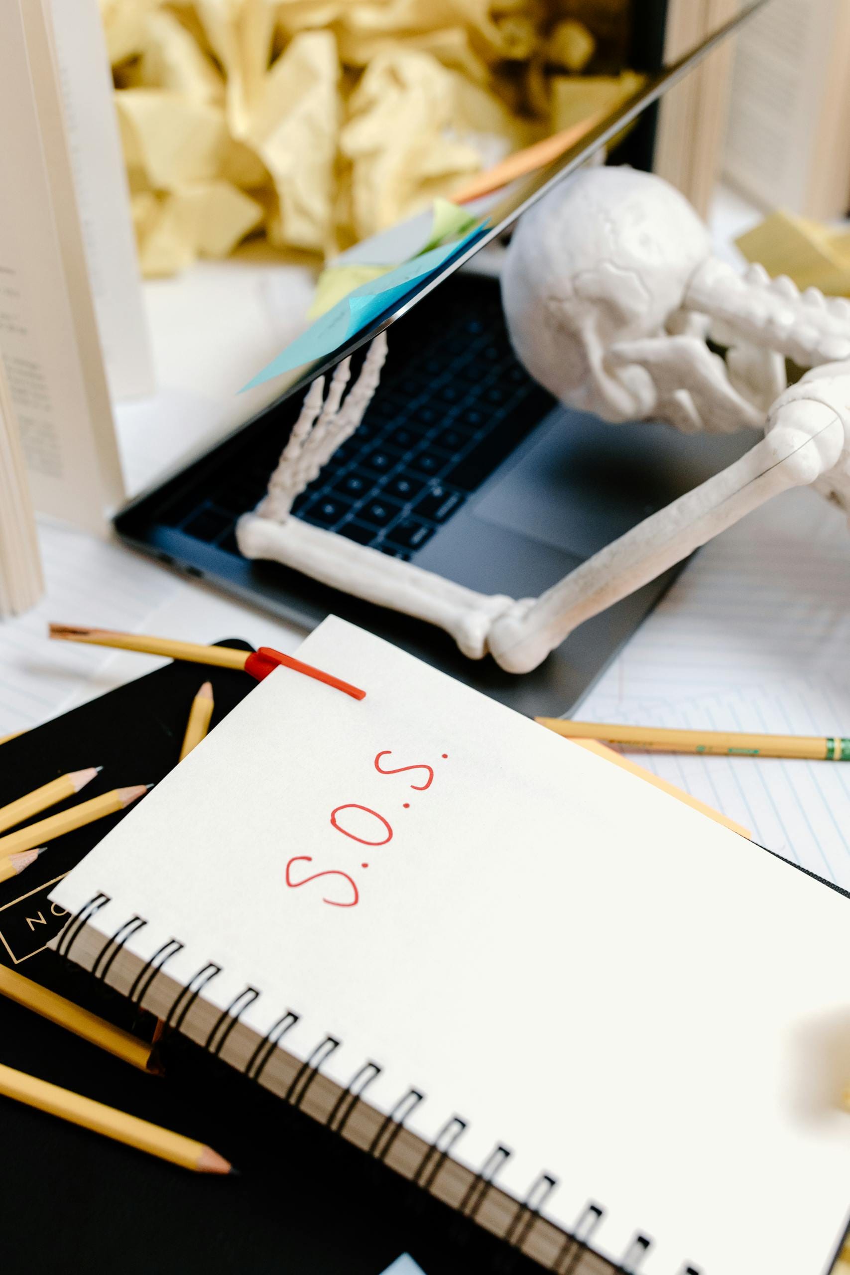 A skeleton figure resting on a laptop with a notepad reading 'SOS' on a cluttered desk.