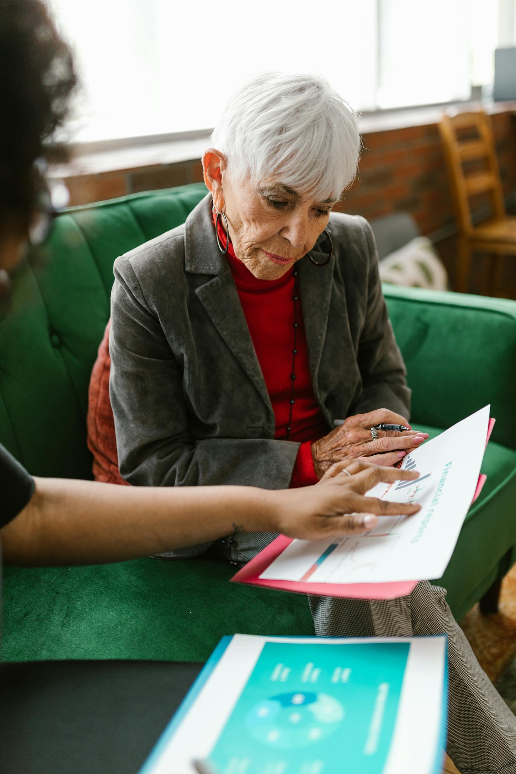 An elderly woman in a grey suit discusses reports with a colleague indoors.